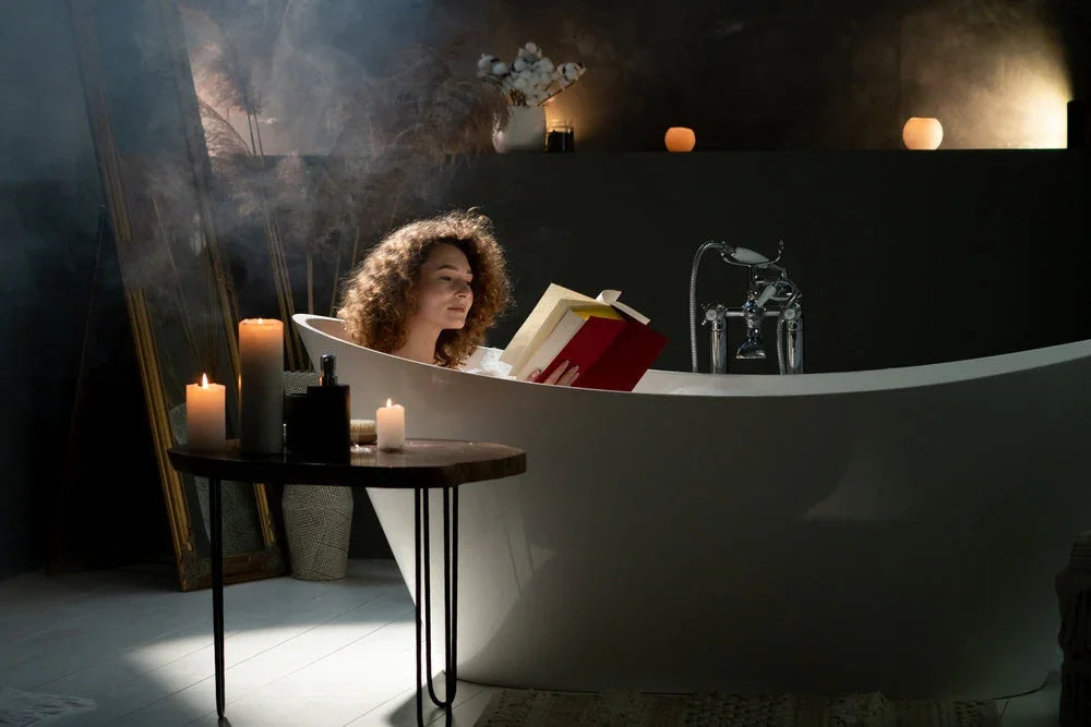Woman with curly hair reading book in white bathtub surrounded by lit candles for relaxation