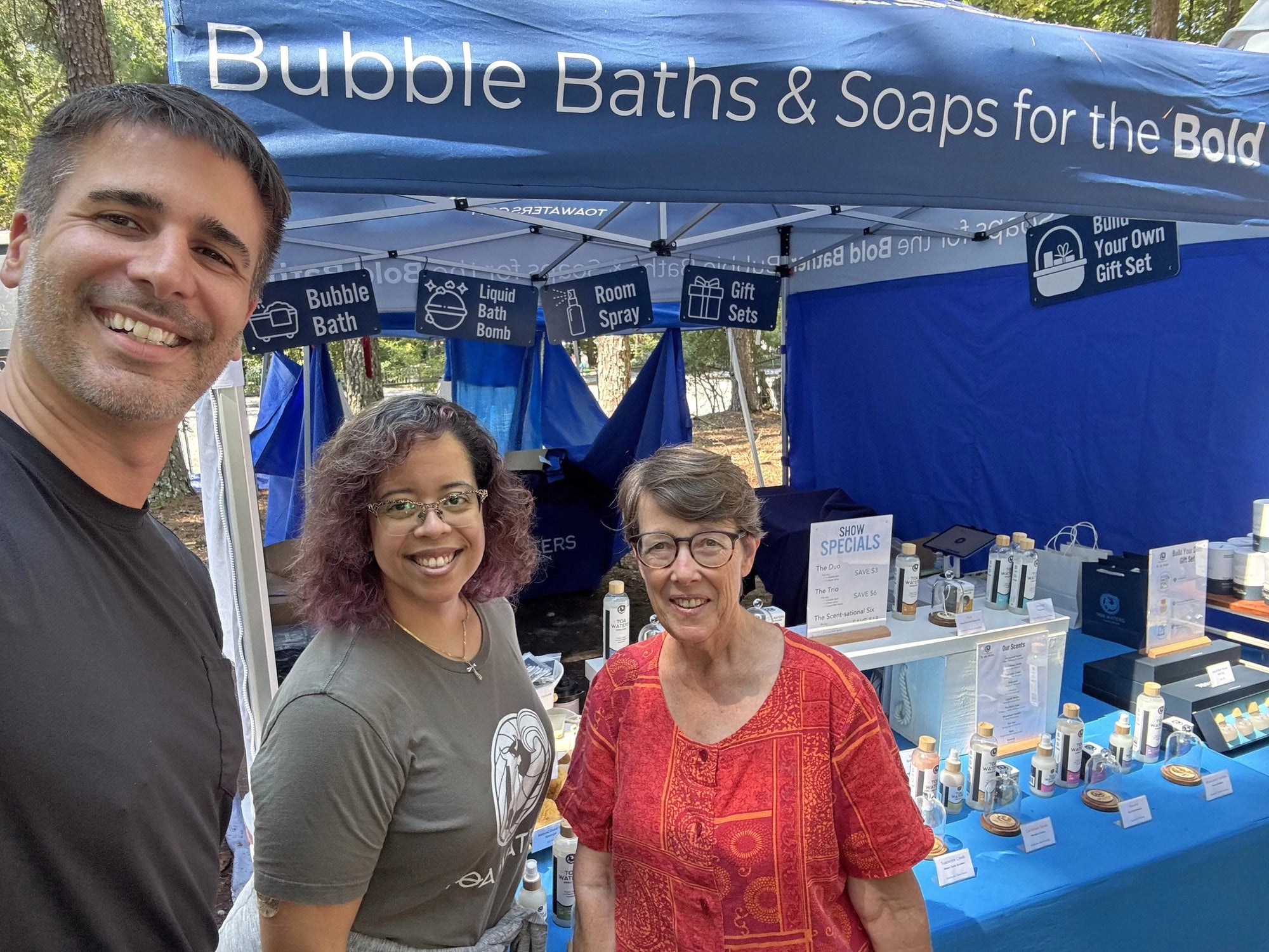 Three people smiling at a market stall with bubble baths, liquid bath bombs, room sprays, and gift sets under a blue canopy.