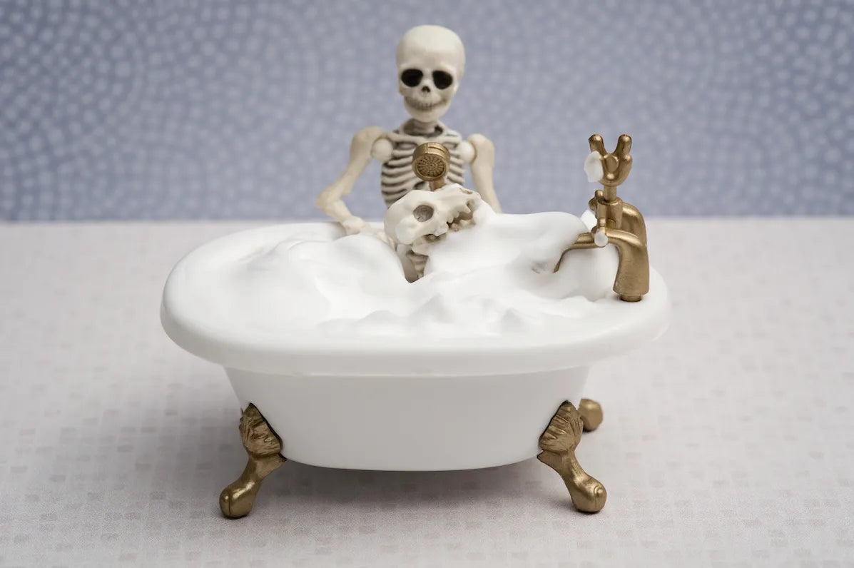 Miniature skeleton figure in a white clawfoot bathtub filled with foam, against a patterned background