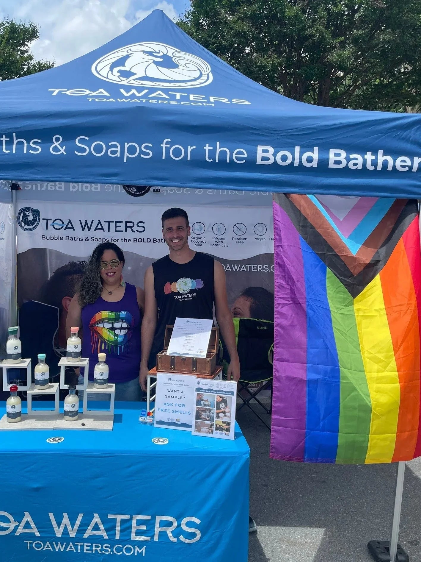 Two people at a TOA Waters booth with bath products and colorful Pride flag at outdoor festival