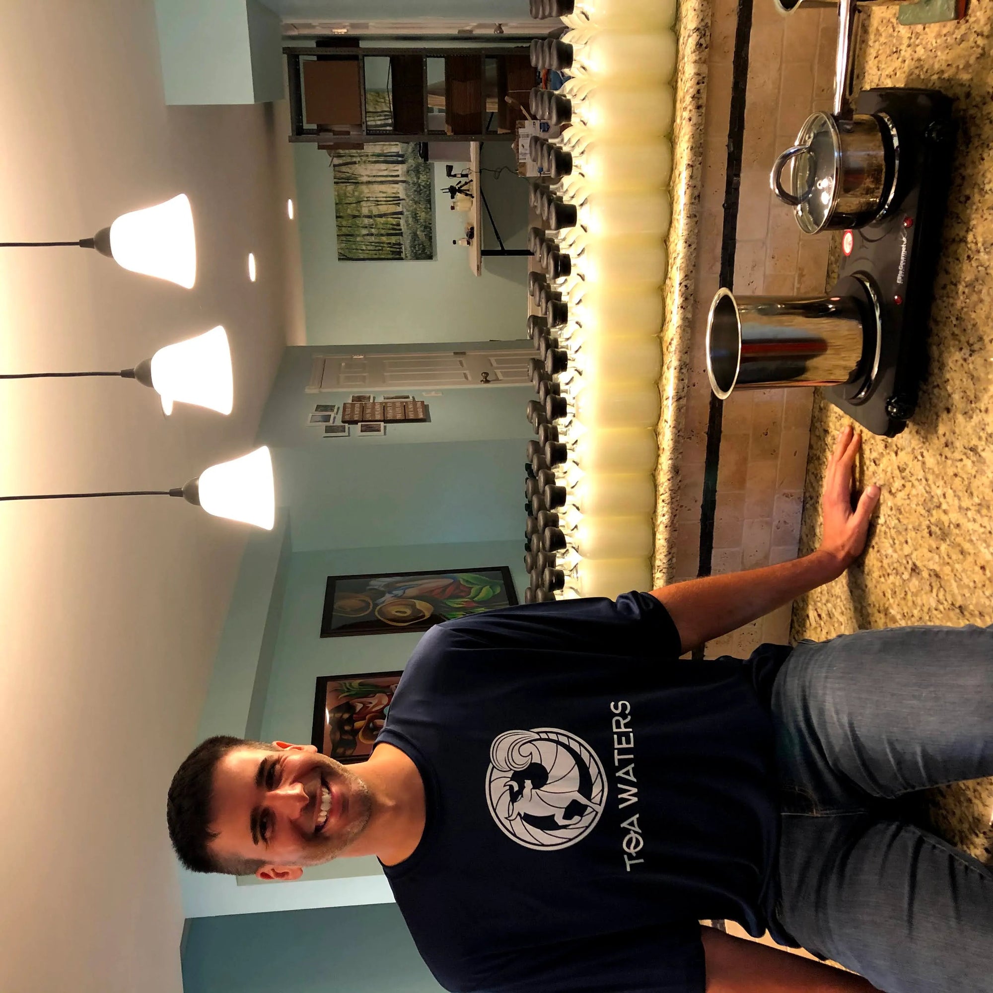 Smiling man wearing TOA Waters shirt standing at kitchen counter with rows of water bottles and stove kettle