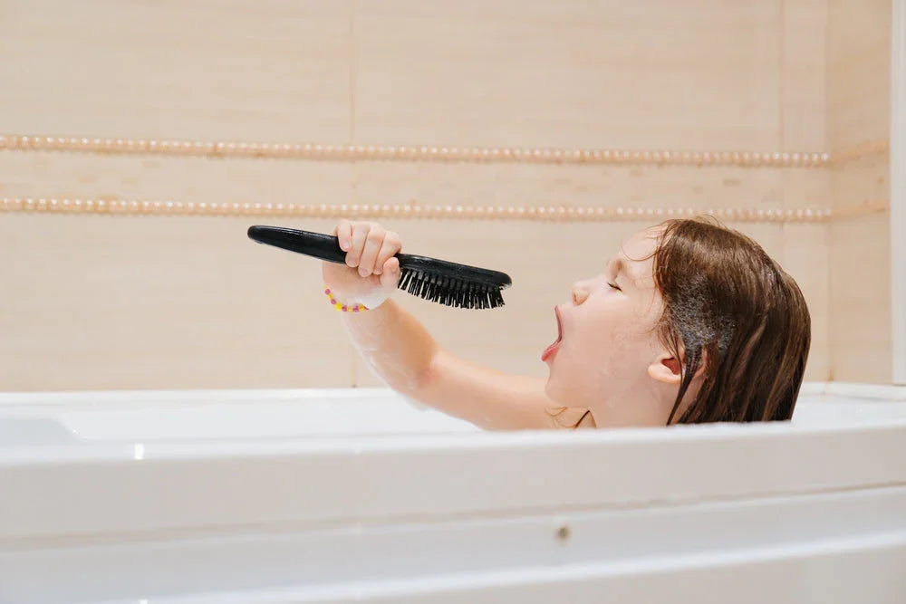 Child singing into a black hairbrush like a microphone while bathing in a white bathtub