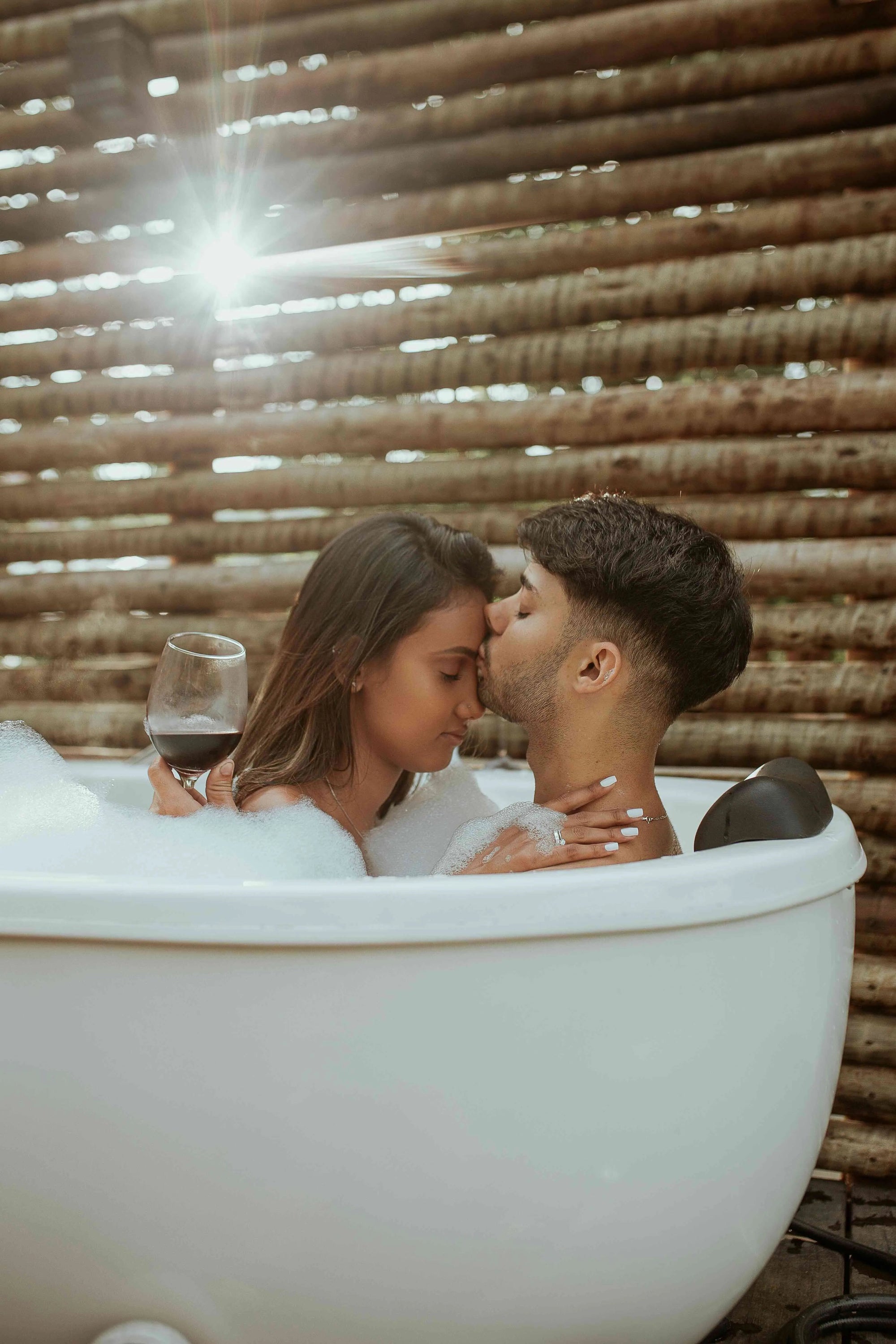 Couple enjoying a warm bubble bath outdoors, woman holding a wine glass, man kissing her forehead