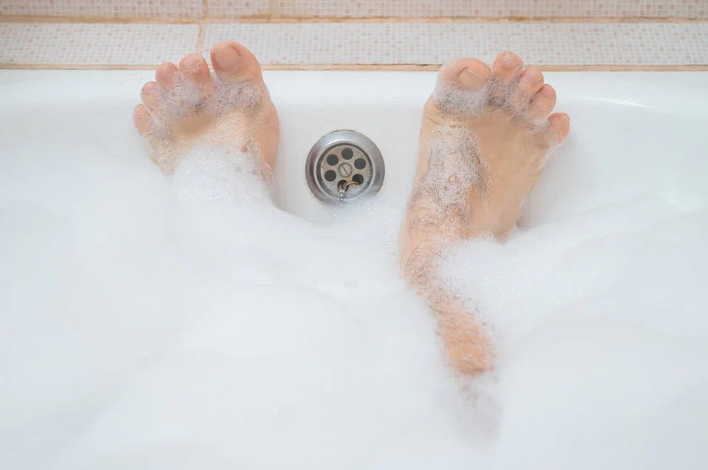 man's feet covered in bubbles in a white bathtub with tiled background and drain visible