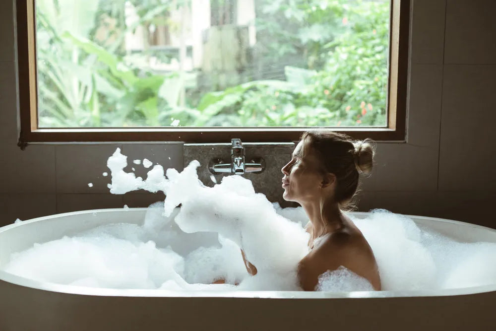 Woman relaxing in foam-filled bathtub near large window with green tropical plants outside