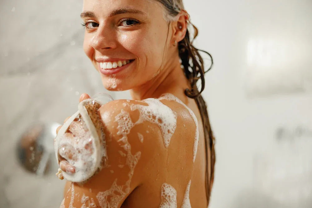 Smiling woman washing her shoulder with soap and exfoliating bath brush in a shower