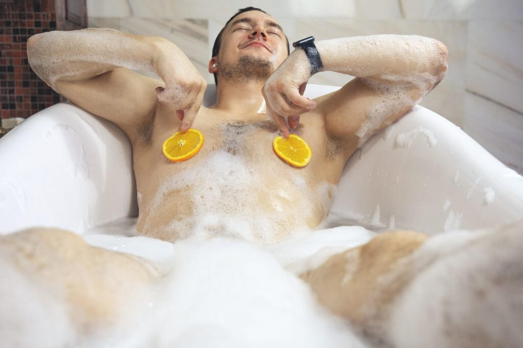 Man holding orange slices over his nipples while taking a bubble bath